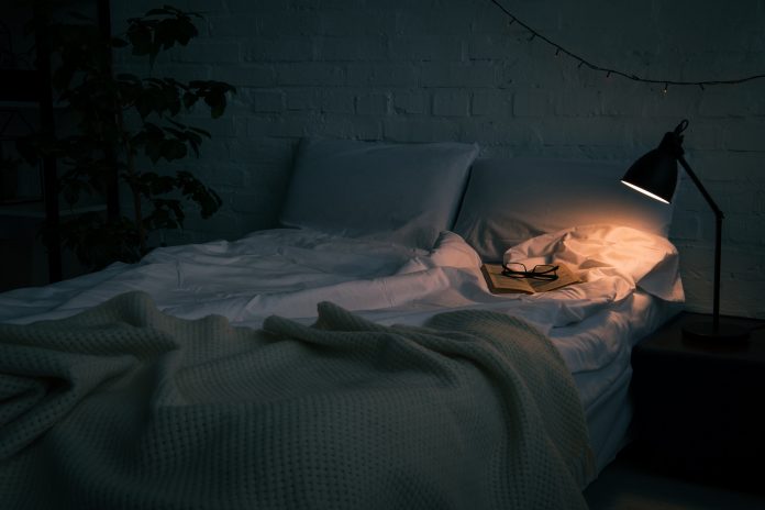 Tmavá ložnice osvětlena světlem ze stolní lampičky. Na posteli leží rozečtená kniha a na ní jsou položeny brýle.