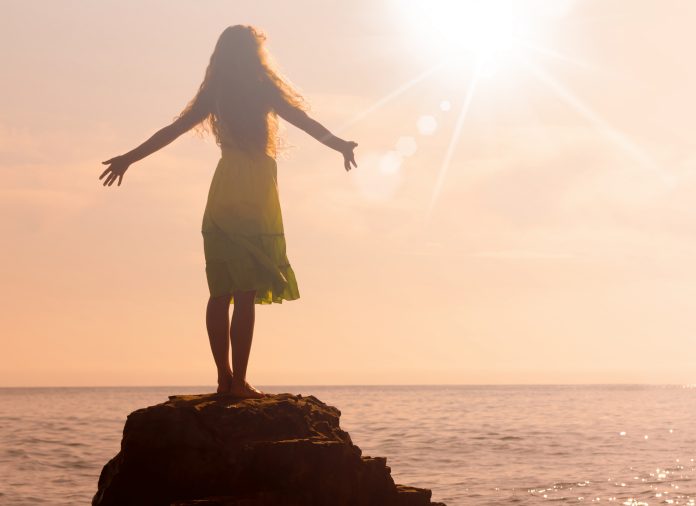 Žena stojí na kameni u moře a roztahuje ruce ke slunci.