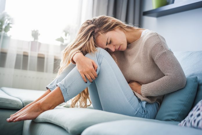 Mladá žena sedící doma na gauči trpí bolestí břicha a křečemi.